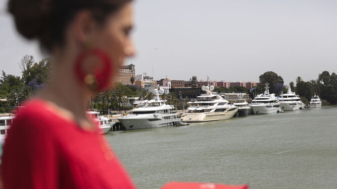 Una mujer, vestida de flamenca, atraviesa el puente de Los Remedios. Al fondo, los siete yates atracados esta semana en el Muelle de las Delicias.