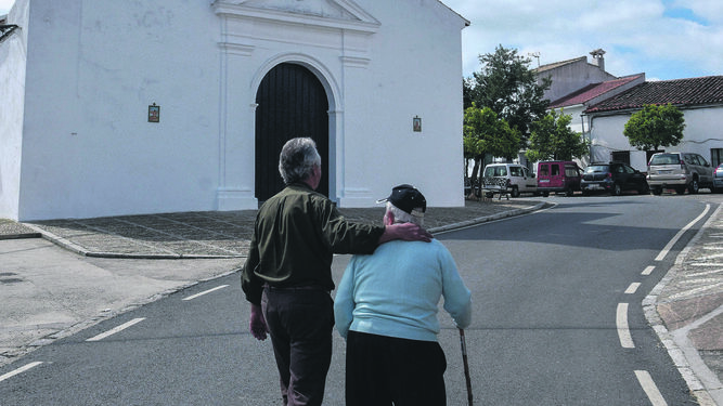 Una pareja de ancianos pasea frente a la iglesia de El Madroño.