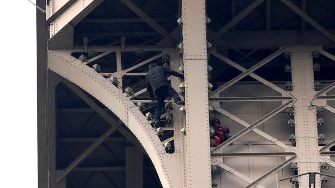 El escalador sube por la estructura de la Torre Eiffel con un bombero vigilante a pocos metros.