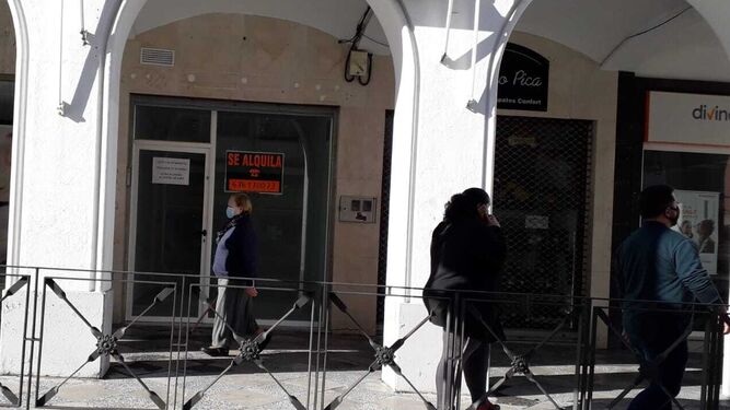 Ciudadanos pasan junto a un negocio cerrado en Esteve en el que luce un cartel que insta a Junta y Ayuntamiento a ponerse de acuerdo porque "estáis matando al centro".